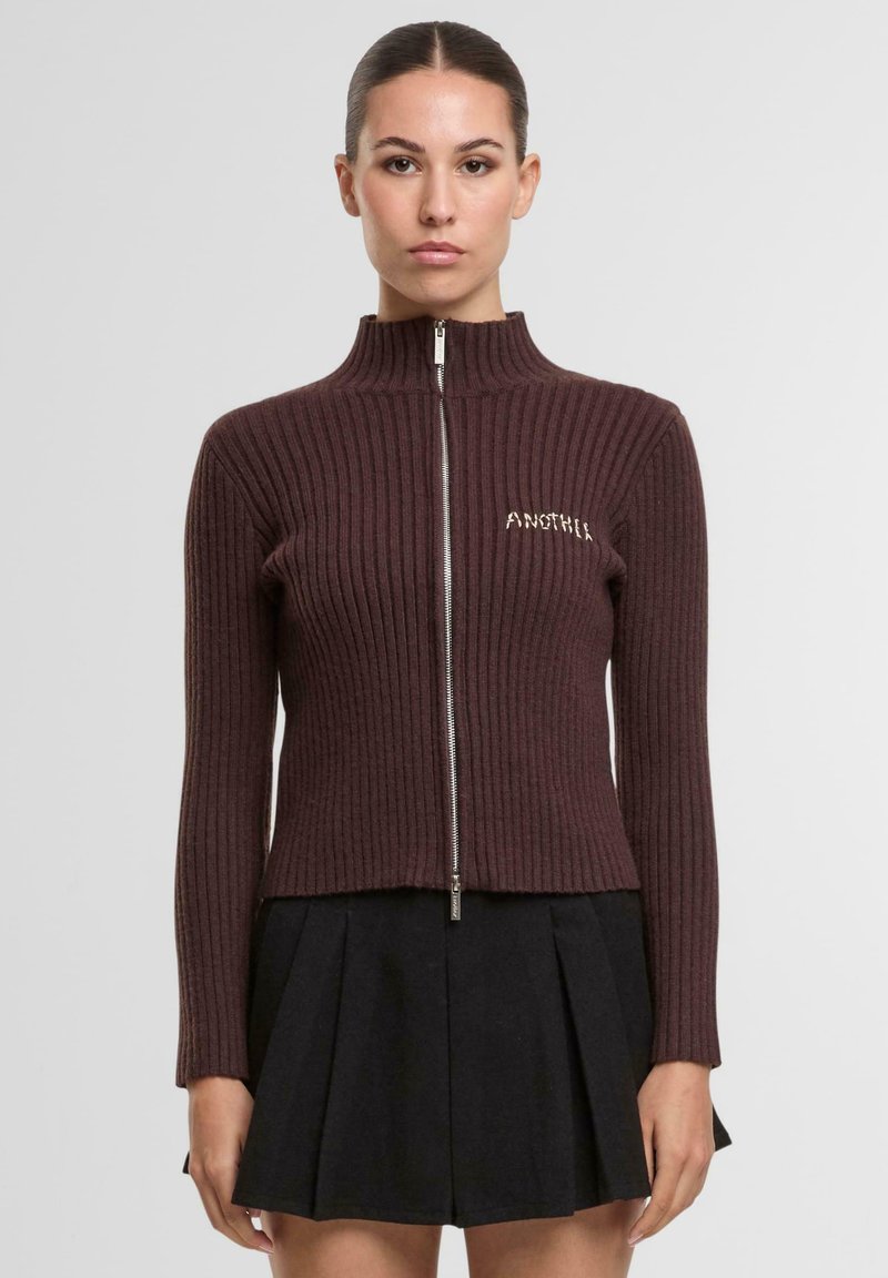 Bordowy prążkowany sweter rozpinany na zamek z wysokim kołnierzem i haftowanym napisem "ANOTHER" na piersi. Noszony z czarną plisowaną spódnicą.