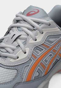 Κοντινή λήψη γκρι και λευκού αθλητικού παπουτσιού με πορτοκαλί λεπτομέρειες και λευκά κορδόνια, με υφασμένα πλαϊνά πάνελ και κόκκινο λογότυπο στη γλώσσα.