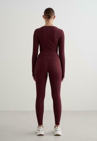 Langärmliges, enganliegendes Oberteil und hochtaillierte Leggings aus burgunderroben, geripptem Material. Die Rückansicht zeigt ein nahtloses Design mit minimalen Details und Sportschuhen.