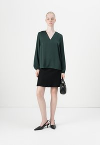 Vrouw staat in een donkergroene blouse met lange mouwen, zwarte rok, zwarte slingback hakken, met een kleine zwarte handtas tegen een witte achtergrond.
