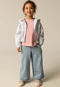 Haut à capuche gris matelassé avec capuche, t-shirt rose à manches courtes et jeans bleu clair à jambes larges. Baskets beiges avec des accents multicolores.