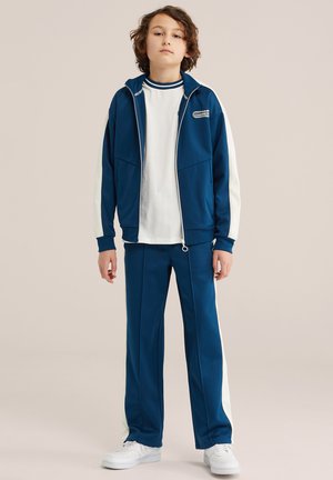 Pois lokkis juustega, kes kannab sinist ja valget dressipluusi ning valgeid vabaajajalatsid, seisab ühevärvilise beeži tausta ees.