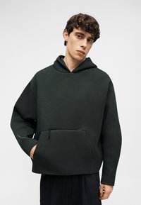 Jeune homme aux cheveux bouclés portant un sweat à capuche ample vert foncé avec poche kangourou et pantalon noir, debout devant un fond clair uni.