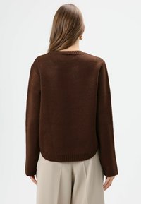 Brauner Strickpullover mit lockerem Schnitt, geripptem Halsausschnitt und langen Ärmeln, kombiniert mit hellbeigen Hosen. Weiche Textur und schlichtes Design.