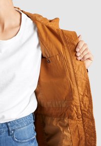 Beżowa kurtka z pikowaną teksturą, pokazująca wewnętrzną kieszeń, noszona na białej koszulce i zestawiona z niebieskimi dżinsami.