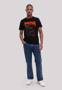 Black t-shirt featuring a serpent graphic in red and green, paired with blue jeans and white sneakers. Model stands against a neutral background.