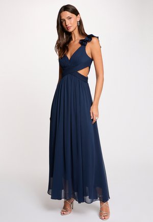 Donna che indossa un abito lungo blu navy con spalline arricciate, scollo a V, spacchi laterali e tacchi alti argentati con cinturini.