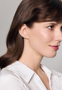 Złote kolczyki w kształcie sztyftów z małym, przezroczystym kryształem w centrum. Wzór jest okrągły, na tle prostej białej koszuli.