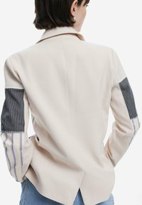 Beige blazer featuring striped fabric accents on sleeves, tailored design, and a vent at the back. Smooth texture, standard collar.