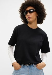 Juodas medvilninis t-shirt su laisvu kirpimu, trumpomis rankovėmis, dėvimas virš baltos ilgomis rankovėmis palaidinės; derinamas su šviesiai mėlynomis džinsinėmis kelnėmis ir saulės akiniais.