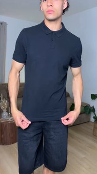 Jeune homme portant un polo noir à manches courtes et un short noir, debout à l'intérieur d'un salon avec des plantes et des meubles.