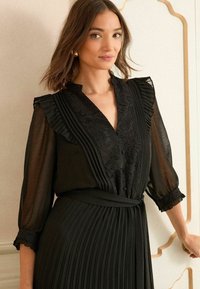 Robe noire avec tissu transparent, détails en dentelle au niveau du col, jupe plissée et accents de manches volantées. Se ferme avec une ceinture.