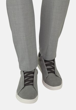 Sneakers in suede grigio con lacci elastici neri, indossate con pantaloni su misura grigio chiaro, viste da davanti a livello della caviglia.