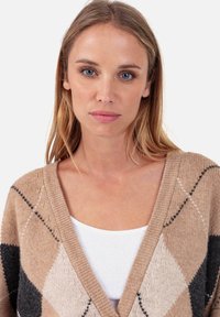 Cardigan en maille argyle beige avec des accents noirs et crème. Design à ouverture frontale, tissu texturé et large col en V. Haut blanc visible à l'intérieur.