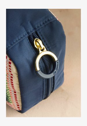 Cipzár sötétkék textil tasakon, arany-fekete kör alakú cipzárhúzóval, oldalán mintás textil panel található.