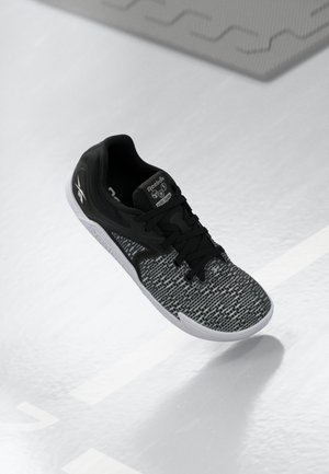 Chaussure de sport Reebok noire et grise avec tige en maille texturée et semelle blanche, montrée flottant au-dessus d'un sol blanc avec des motifs subtils.