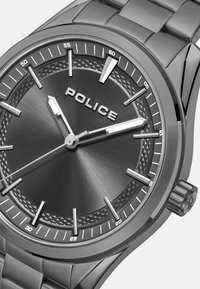 Srebrna ura iz nerjavečega jekla z teksturirano črno številčnico, indeksnimi oznakami in vidno prikazanim imenom blagovne znamke "POLICE".
