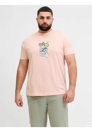 Homme avec barbe portant un t-shirt rose clair avec un graphique de boisson et de main, et un short vert clair, debout devant un fond uni.