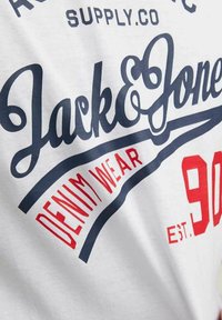 Tissu blanc avec texte bleu marine et rouge indiquant « Jack & Jones Denim Wear Est. 90 » en polices grasses stylisées sur un vêtement décontracté.