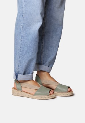 Green leather espadrille sandals with a woven jute sole and elastic straps, paired with rolled-up light blue denim jeans.