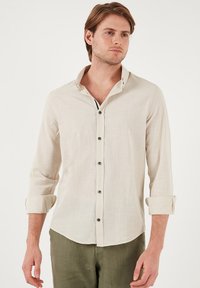 Camisa de lino beige con cuello abotonado, mangas largas remangadas, seis botones en el frente y una tela texturizada.