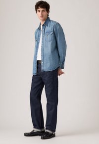 Uomo con camicia di denim azzurro chiaro sopra una t-shirt bianca, jeans blu scuro con risvolti, mocassini in pelle nera, in piedi contro uno sfondo uniforme.