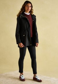 Czarna wodoodporna kurtka z ściągaczem w talii, dwoma przednimi kieszeniami, noszona na bordowym swetrze i czarnych spodniach. Brązowe i niebieskie trampki uzupełniają wygląd.