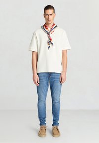 Camicia a maniche corte color crema, jeans in denim blu, mocassini beige e una sciarpa multicolore con motivo annodata intorno al collo. Look semplice e casual.