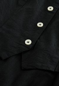 Chemise en tissu noir avec une surface texturée, présentant des boutons écrèmes avec des découpes en forme d'étoile et une apparence légèrement froissée.