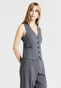 Gilet gris rayé avec un col en V, comprenant quatre boutons noirs et une coupe ajustée. Tissu lisse et coupe élégante, associé à un pantalon assorti.