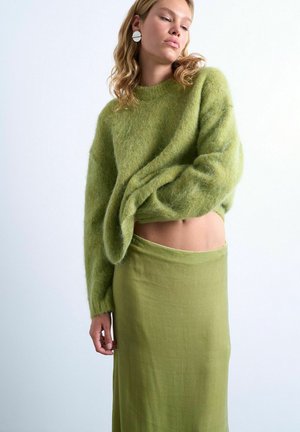 Donna che si tira su un maglione verde peloso, indossa una gonna verde abbinata e orecchini bianchi a cerchio, davanti a uno sfondo chiaro e neutro.