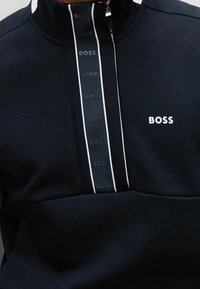Sudadera con capucha en color navy, con cuello alto, franjas blancas en los costados, media cremallera en la parte delantera, logo impreso en blanco en el pecho y textura de tela suave.