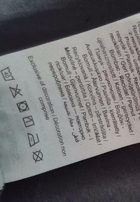 Štítok s viacjazyčným textom, symbolmi starostlivosti a ikonami recyklácie, vytlačený na jemnom textilnom materiáli svetlej farby.