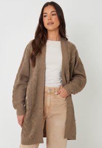 Chaleco de punto en marrón claro con patrones de trenzado, de frente abierto y de manga larga. Lleva puesto sobre una blusa blanca y combinado con pantalones beige.