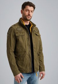 Veste vert olive avec fermeture éclair, deux poches poitrine et doublure intérieure jaune. Comprend des empiècements aux épaules et un design décontracté.