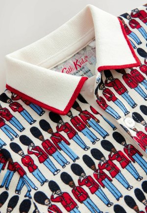 Pikétröja i ett texturerat krämfärgat tyg med ett upprepande mönster av soldater i röda uniformer och blå byxor, konturerade i svart. Röda krage-accenter.