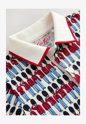 Cămașă polo dintr-un material texturat de crem, cu un model repetitiv de soldați în uniforme roșii și pantaloni albaștri, conturați în negru. Accente roșii la guler.