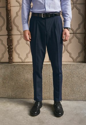 Man wearing navy blue pleated trousers, black belt, black dress shoes, striped blue dress shirt, and wristwatch standing indoors.