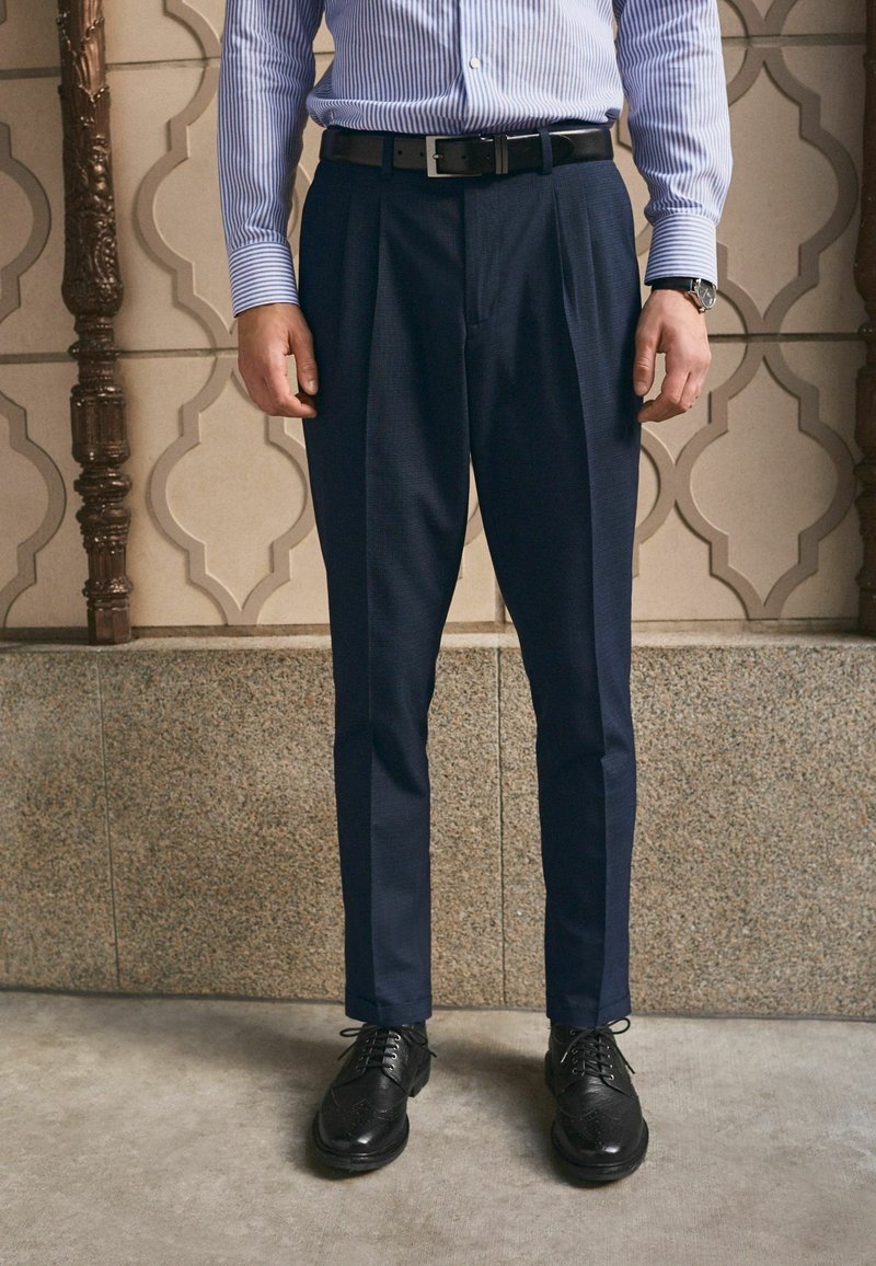 Man wearing navy blue pleated trousers, black belt, black dress shoes, striped blue dress shirt, and wristwatch standing indoors.