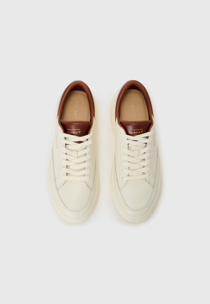 Zapatillas de piel blancas con un acento en el talón de piel marrón, cordones planos y una textura suave. La marca aparece en la lengua.