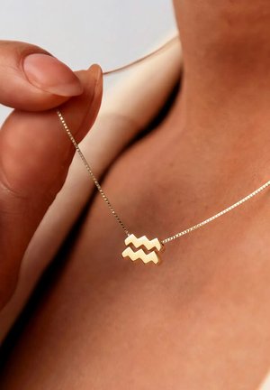 Persoon die een gouden ketting vasthoudt met een zigzaggend Waterman sterrenbeeldhanger tegen blootgestelde huid en witte kleding.