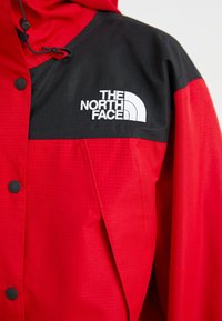 Rote und schwarze Jacke aus glattem, leichtem Stoff; mit Kapuze, Druckknöpfen und gesticktem Logo auf der Brust.