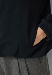 Czarny pulower o miękkiej teksturze, charakteryzujący się luźnym krojem i ściągaczami przy rękawach, zestawiony z szarymi plisowanymi spodniami. Z boku widoczna kieszeń.