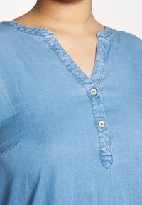 Světle modrá džínová košile s výstřihem do V a dvěma malými bílými knoflíčky poblíž límce na horní části těla člověka.