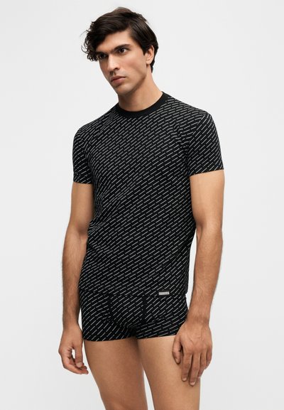 Homme portant une chemise noire à manches courtes assortie et un boxer avec un petit motif en diagonale de texte blanc sur un fond uni.