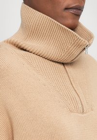 Gros plan d'une personne portant un pull en maille côtelée beige avec un col montant et une fermeture éclair argentée partiellement ouverte.