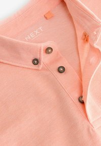 Polo de manga corta en melocotón claro con un tejido texturizado, tres botones oscuros y una etiqueta de tela etiquetada "NEXT" en el interior del cuello.