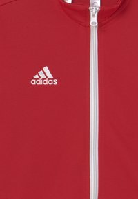 Chaqueta deportiva roja hecha de tela texturizada con cierre de cremallera blanco. Cuenta con un pequeño logo blanco de Adidas en el lado izquierdo.