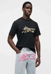 Μαύρο βαμβακερό μπλουζάκι με γραφικό τιγρέ καμουφλάζ. Ανοιχτό μπλε τζιν παντελόνι με ροζ περίγραμμα παρόμοιας τίγρης στον μηρό.