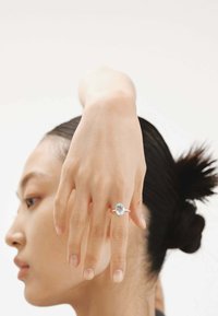 Anillo de plata con una gran piedra de aguamarina ovalada, montada sobre una banda delgada. Mano posada elegantemente contra un fondo neutro.